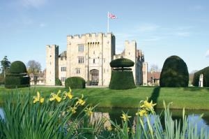 Ein majestätisches Schloss im englischen Stil steht im Hintergrund, mit steinernen Mauern und Türmen. Im Vordergrund ist ein gepflegter Garten zu sehen, mit kunstvoll geschnittenen Buchsbäumen in runden Formen. Ein kleiner Teich spiegelt den blauen Himmel wider. Frühlingsblumen, darunter gelbe Narzissen und weiße Schneeglöckchen, blühen im Vordergrund zwischen grünen Gräsern. Eine britische Flagge weht auf einem der Schlosstürme und unterstreicht die historische Atmosphäre der Szene.