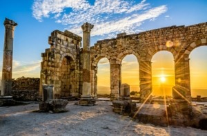 Sonnenuntergang an einer antiken römischen Ruine. Im Vordergrund stehen Säulen und Steinmauern mit Bogenöffnungen. Durch die Bögen scheint die untergehende Sonne, die den Himmel in warme Goldtöne taucht. Weiße Wolken am blauen Himmel bilden einen malerischen Kontrast zu den alten Steinstrukturen.