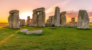 Antike Steinkreisformation von Stonehenge bei Sonnenuntergang. Massive, aufrecht stehende Steine bilden einen kreisförmigen Aufbau auf einer grünen Wiese. Der Himmel ist in warme Orange- und Rosatöne getaucht, die einen dramatischen Hintergrund für die mysteriöse prähistorische Stätte bilden.