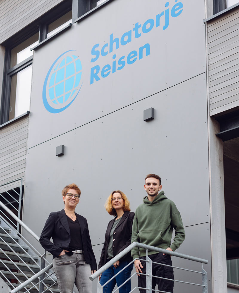 Drei Mitarbeitende von Schatorjé Reisen stehen vor dem Firmengebäude mit Logo.