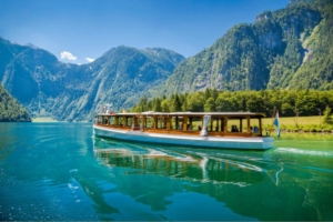 Dieses Bild zeigt eine atemberaubende Szene am Königssee in Bayern, einem der schönsten Alpenseen Deutschlands. Im Vordergrund sieht man ein traditionelles Elektroboot, das für Touristenfahrten auf dem See verwendet wird. Das Boot ist lang und schmal, typisch für die Königssee-Boote, und bietet Platz für viele Passagiere unter einem Dach zum Schutz vor Sonne oder Regen. Der See selbst ist von einer beeindruckenden tiefblauen Farbe und spiegelt den klaren Himmel und die umliegenden Berge wider. Das Wasser ist so klar, dass man fast bis auf den Grund sehen kann. Im Hintergrund erheben sich majestätische Alpen mit steilen, bewaldeten Hängen, die direkt bis zum Seeufer reichen. Die Berge sind teilweise von Wolken umhüllt, was der Szene eine mystische Atmosphäre verleiht. Die Landschaft strahlt eine unberührte, natürliche Schönheit aus. Die Kombination aus dem klaren See, den imposanten Bergen und dem traditionellen Boot schafft ein Bild von zeitloser Ruhe und Erhabenheit. Diese Szene ist typisch für die Berchtesgadener Region und zeigt, warum der Königssee ein beliebtes Reiseziel für Naturliebhaber und Touristen ist. Sie vermittelt ein Gefühl von Frieden und Harmonie zwischen Mensch und Natur.