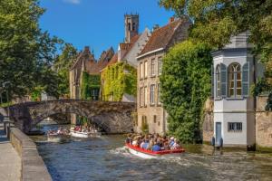 Dieses Bild zeigt eine malerische Szene aus einer historischen europäischen Stadt, höchstwahrscheinlich Brügge in Belgien. Im Vordergrund sieht man einen Kanal, auf dem ein rotes Touristenboot mit mehreren Passagieren fährt. Der Kanal wird von einer alten Steinbrücke überspannt, die den Charme der mittelalterlichen Architektur unterstreicht. Entlang des Kanals stehen wunderschöne alte Gebäude mit charakteristischen Giebeldächern und Fassaden aus Backstein. Einige der Häuser sind mit Efeu oder anderen Kletterpflanzen bewachsen, was dem Bild eine besonders romantische Note verleiht. Die Bäume am Ufer des Kanals spenden Schatten und verstärken den idyllischen Eindruck der Szene. Der blaue Himmel mit einigen weißen Wolken bildet einen schönen Kontrast zu den warmen Farbtönen der Gebäude. Diese Aufnahme vermittelt perfekt die Atmosphäre einer historischen europäischen Stadt mit ihren Wasserstraßen, alten Brücken und gut erhaltenen mittelalterlichen Gebäuden. Es ist ein Bild, das Besucher dazu einlädt, die Geschichte und Schönheit der Stadt zu erkunden und die friedliche Atmosphäre zu genießen.