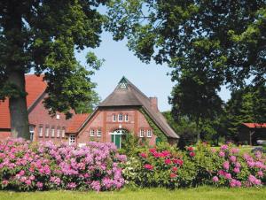 Dieses Bild zeigt ein charmantes ländliches Gebäude, vermutlich ein altes Bauernhaus oder Cottage. Das Haus hat ein steiles, mit Ziegeln gedecktes Dach und scheint aus Ziegelsteinen oder einem ähnlichen traditionellen Material gebaut zu sein. Im Vordergrund des Bildes ist ein üppiger Garten zu sehen. Besonders auffällig ist ein großer blühender Busch oder Baum mit rosa Blüten, der einen schönen Kontrast zum roten Dach und den grünen Bäumen im Hintergrund bildet. Die Umgebung des Hauses ist sehr grün und natürlich. Große Bäume rahmen das Haus ein und schaffen eine friedliche, idyllische Atmosphäre. Der Himmel ist blau mit einigen weißen Wolken, was auf einen schönen Sommertag hindeutet. Insgesamt vermittelt das Bild den Eindruck eines malerischen, ländlichen Refugiums - ein Ort, der Ruhe und Naturverbundenheit ausstrahlt. 