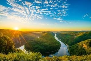 Dieses Bild zeigt eine atemberaubende Landschaftsaufnahme eines Flusses, der sich durch eine grüne, bewaldete Landschaft schlängelt. Im Vordergrund sehen wir den Fluss, der eine markante S-Kurve bildet. Das Wasser spiegelt den Himmel wider und glitzert im Sonnenlicht. Die Ufer des Flusses sind dicht mit Bäumen und Vegetation bewachsen, was der Szene eine üppige, natürliche Atmosphäre verleiht. Die Landschaft erstreckt sich weit in den Hintergrund und zeigt sanfte Hügel und Täler, alle bedeckt mit dichtem, grünem Wald. Die Baumkronen bilden ein nahezu ununterbrochenes Blätterdach, das sich bis zum Horizont erstreckt. Am Himmel sieht man ein beeindruckendes Wolkenmuster. Kleine, flauschige Wolken bedecken den größten Teil des Himmels und bilden einen interessanten Kontrast zum klaren Blau dahinter. Die Sonne geht gerade auf oder unter und taucht die Szene in ein warmes, goldenes Licht. Die Sonnenstrahlen brechen durch die Wolken und beleuchten Teile der Landschaft, was dramatische Licht- und Schatteneffekte erzeugt. Insgesamt vermittelt das Bild einen Eindruck von unberührter Natur, Ruhe und Schönheit. Es ist eine Szene, die Ehrfurcht und Bewunderung für die natürliche Welt hervorruft.