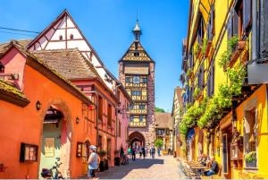 Dieses Bild zeigt eine malerische Straßenszene in einer historischen europäischen Altstadt, höchstwahrscheinlich in Deutschland oder einer ähnlichen Region. Im Vordergrund sehen wir eine gepflasterte Gasse, die von bunten, traditionellen Fachwerkhäusern gesäumt wird. Die Häuser haben charakteristische Holzbalken, die sich durch die farbigen Fassaden ziehen, und sind in warmen Tönen wie Gelb, Orange und Rot gestrichen. Einige der Gebäude haben dekorative Erker und Blumenkästen an den Fenstern, was dem Ort einen charmanten, einladenden Charakter verleiht. In der Mitte der Gasse steht ein markanter Uhrenturm, der vermutlich zu einem historischen Rathaus oder einem ähnlichen öffentlichen Gebäude gehört. Der Turm ragt über die umliegenden Häuser hinaus und bildet einen zentralen Blickfang in der Szene. Die Straße selbst ist autofrei und scheint eine Fußgängerzone zu sein. Einige Menschen sind zu sehen, die die Gasse entlangschlendern, was auf eine beliebte touristische Gegend hindeutet. Der Himmel ist blau mit einigen weißen Wolken, was eine angenehme, sonnige Atmosphäre vermittelt. Insgesamt präsentiert das Bild eine typische Ansicht einer gut erhaltenen, historischen Altstadt in Mitteleuropa. Es strahlt eine Atmosphäre von Gemütlichkeit, Geschichte und Kultur aus und lädt den Betrachter ein, sich vorzustellen, wie es wäre, durch diese charmanten Straßen zu spazieren.