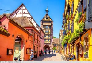 Dieses Bild zeigt eine malerische Straßenszene in einer historischen europäischen Altstadt, höchstwahrscheinlich in Deutschland oder einer ähnlichen Region.

Im Vordergrund sehen wir eine gepflasterte Gasse, die von bunten, traditionellen Fachwerkhäusern gesäumt wird. Die Häuser haben charakteristische Holzbalken, die sich durch die farbigen Fassaden ziehen, und sind in warmen Tönen wie Gelb, Orange und Rot gestrichen. Einige der Gebäude haben dekorative Erker und Blumenkästen an den Fenstern, was dem Ort einen charmanten, einladenden Charakter verleiht.

In der Mitte der Gasse steht ein markanter Uhrenturm, der vermutlich zu einem historischen Rathaus oder einem ähnlichen öffentlichen Gebäude gehört. Der Turm ragt über die umliegenden Häuser hinaus und bildet einen zentralen Blickfang in der Szene.

Die Straße selbst ist autofrei und scheint eine Fußgängerzone zu sein. Einige Menschen sind zu sehen, die die Gasse entlangschlendern, was auf eine beliebte touristische Gegend hindeutet.

Der Himmel ist blau mit einigen weißen Wolken, was eine angenehme, sonnige Atmosphäre vermittelt.

Insgesamt präsentiert das Bild eine typische Ansicht einer gut erhaltenen, historischen Altstadt in Mitteleuropa. Es strahlt eine Atmosphäre von Gemütlichkeit, Geschichte und Kultur aus und lädt den Betrachter ein, sich vorzustellen, wie es wäre, durch diese charmanten Straßen zu spazieren.