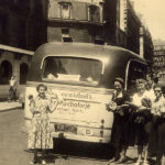 Historische Aufnahme eines alten Busses. Davor steht eine Gruppe Menschen