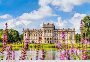 Dieses Bild zeigt das prachtvolle Schweriner Schloss, ein beeindruckendes Beispiel für romantische Historismus-Architektur in Deutschland.

Das Schloss liegt malerisch auf einer Insel im Schweriner See. Seine zahlreichen Türme, Kuppeln und Erker spiegeln sich im ruhigen Wasser des Sees und schaffen so eine fast märchenhafte Atmosphäre. Die Architektur des Schlosses ist eine Mischung aus verschiedenen Stilen, darunter neugotische und Renaissance-Elemente, die ihm ein einzigartiges und majestätisches Aussehen verleihen.

Im Vordergrund sieht man einen Teil der gepflegten Gartenanlagen, die das Schloss umgeben. Grüne Rasenflächen und sorgfältig gestaltete Blumenbeete ergänzen die Schönheit des Gebäudes.

Der Himmel über dem Schloss ist von weißen, flauschigen Wolken bedeckt, die sich ebenfalls im Wasser spiegeln und so die Szene noch malerischer gestalten.

Das Schweriner Schloss, oft als "Neuschwanstein des Nordens" bezeichnet, ist nicht nur ein bedeutendes architektonisches Denkmal, sondern auch Sitz des Landtags von Mecklenburg-Vorpommern. Es ist ein beliebtes Touristenziel und ein Symbol für die reiche Geschichte und Kultur der Region.

Dieses Bild vermittelt einen Eindruck von der Pracht und Schönheit dieses außergewöhnlichen Baudenkmals und lädt den Betrachter ein, sich die Geschichte und die Geschichten vorzustellen, die sich hinter seinen Mauern verbergen.